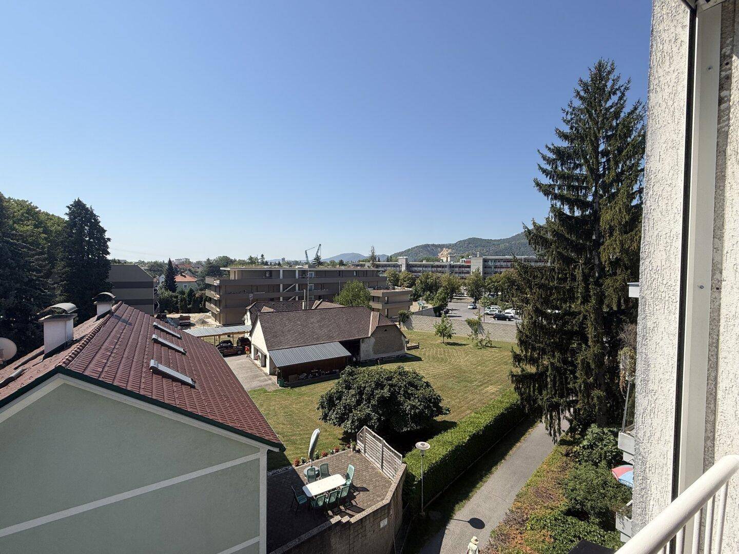 Weitläufiger Ausblick vom Balkon über Dächer, grüne Flächen und eine bergige Landschaft.