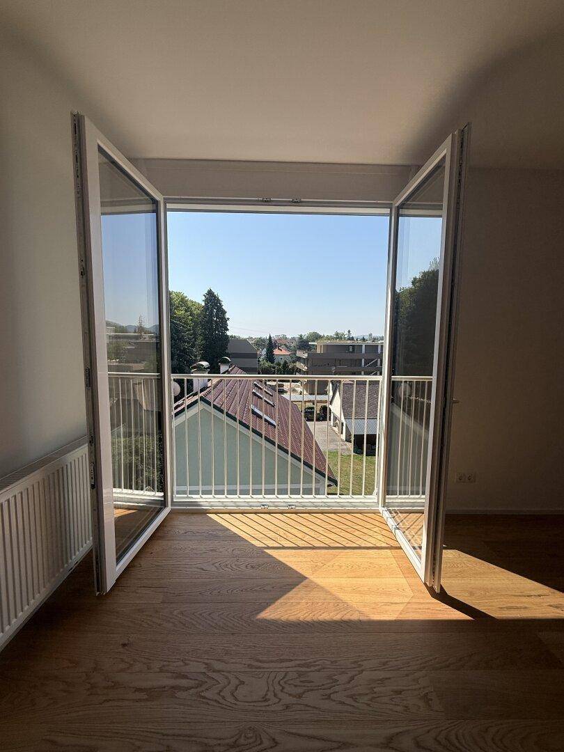 Offene Balkontüren bieten einen weiten Blick über Dächer und grüne Umgebung, viel Sonnenlicht.