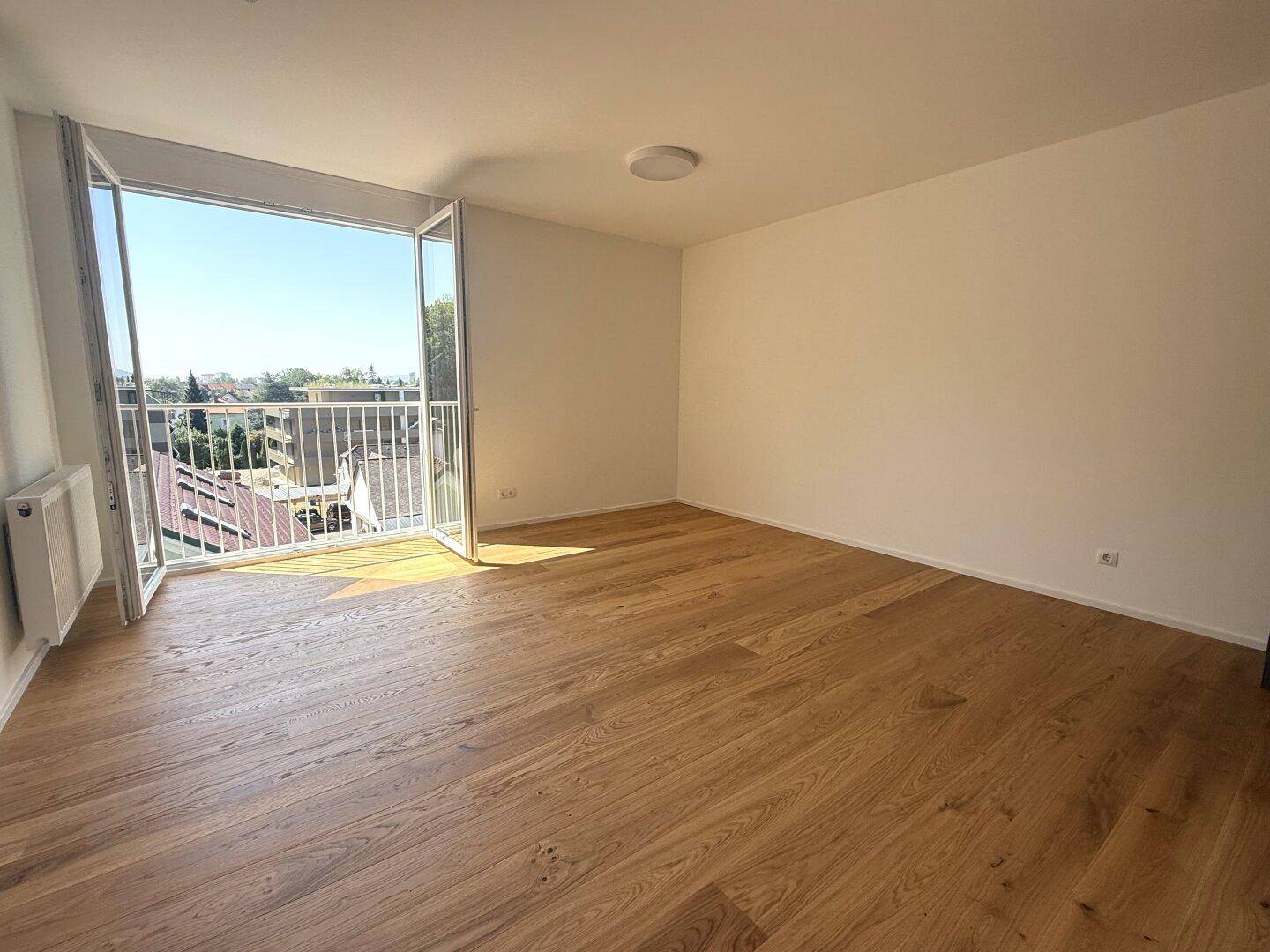 Helles Zimmer mit Holzboden und weit geöffneten Balkontüren, die viel Licht hereinlassen.