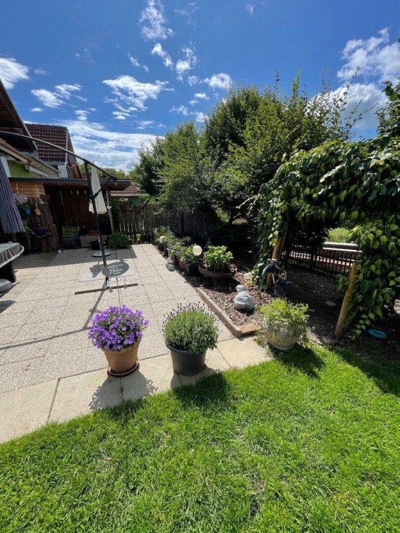 Sonniger Gartenbereich mit gepflasterter Terrasse, Blumen und üppiger Bepflanzung.
