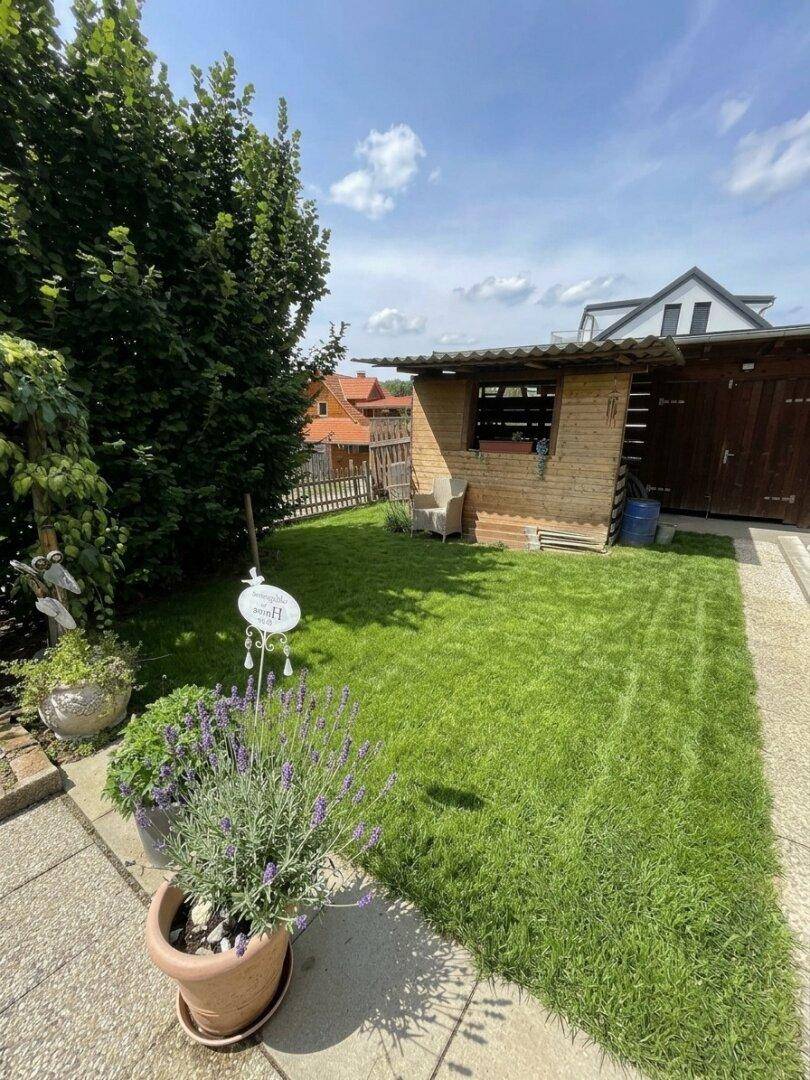 Großer Garten mit Rasenfläche, Gartenhaus und hohen Bäumen im Hintergrund.