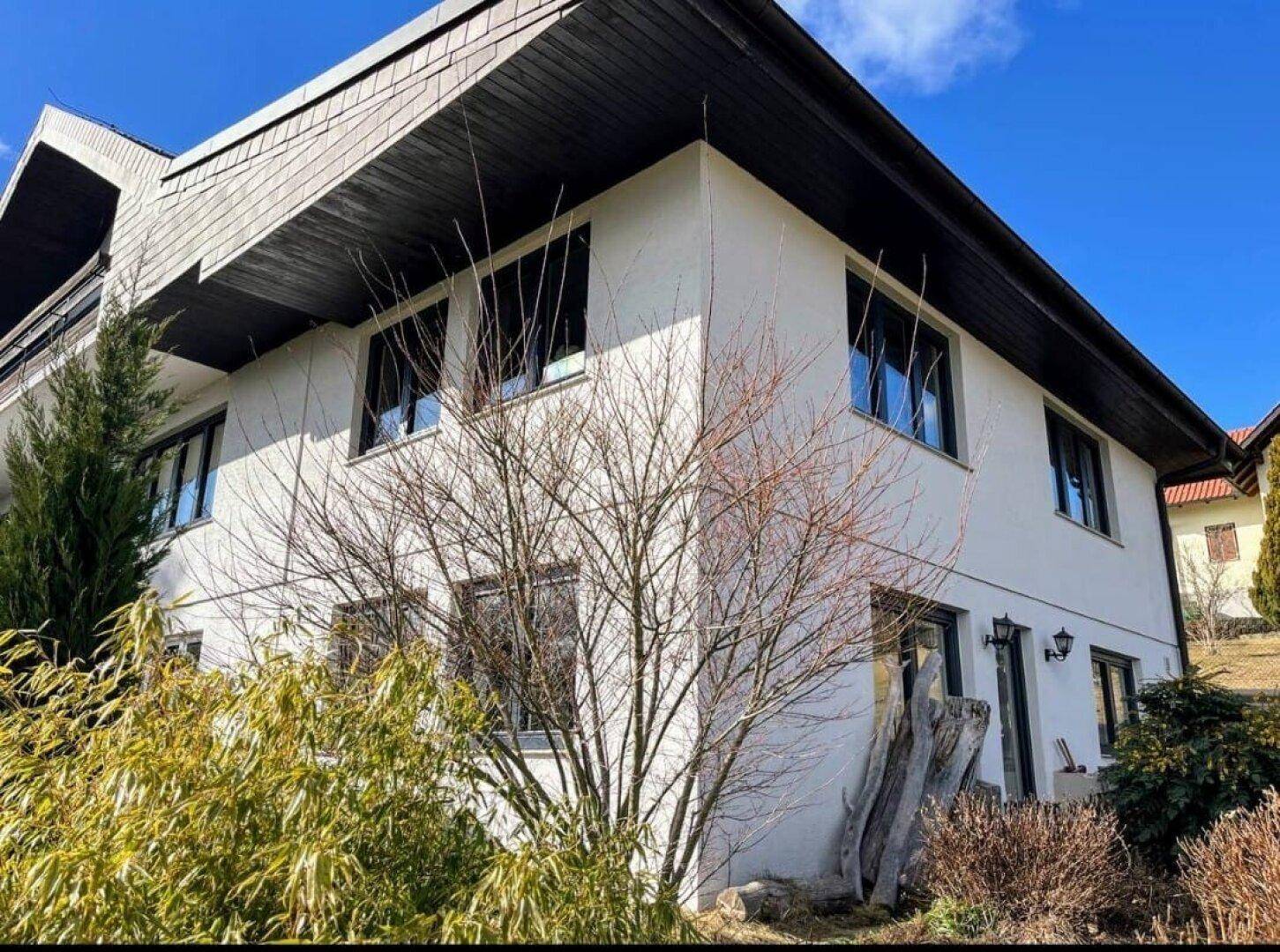 Weiße Hausfassade mit dunklem Dach und Fenstern, umgeben von Bäumen.
