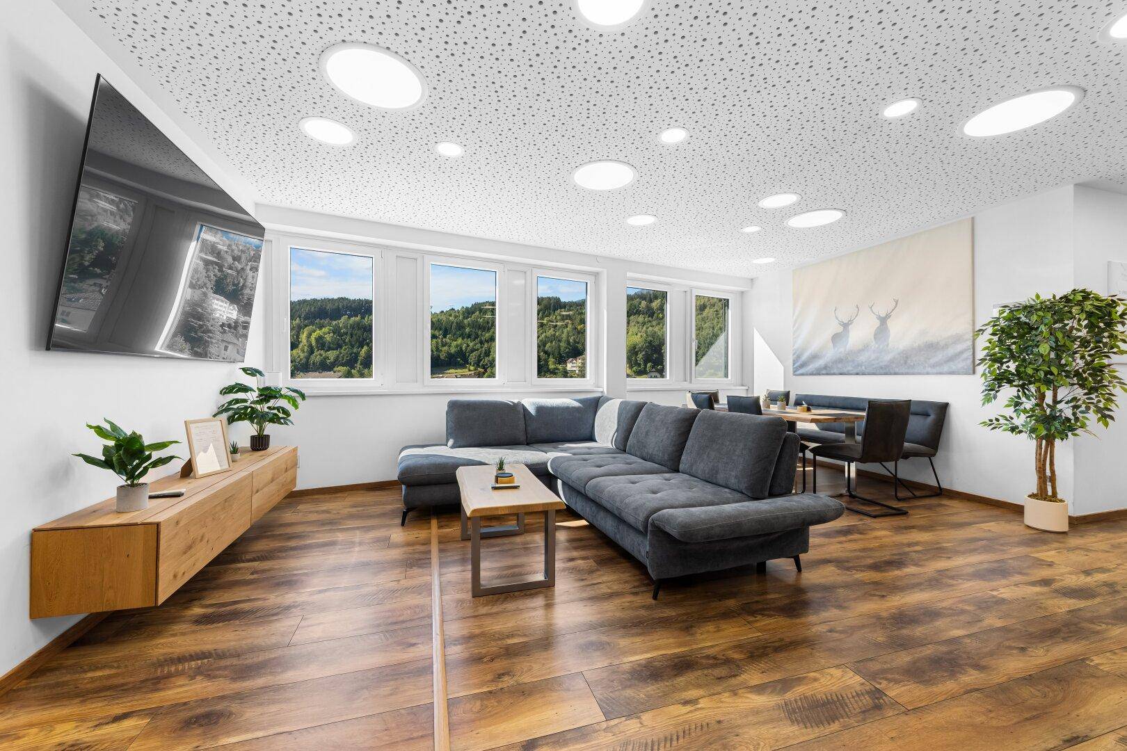 Geräumiges Wohn- und Esszimmer mit gemütlichem Sofa, Holzboden und Blick auf die bewaldeten Hügel.