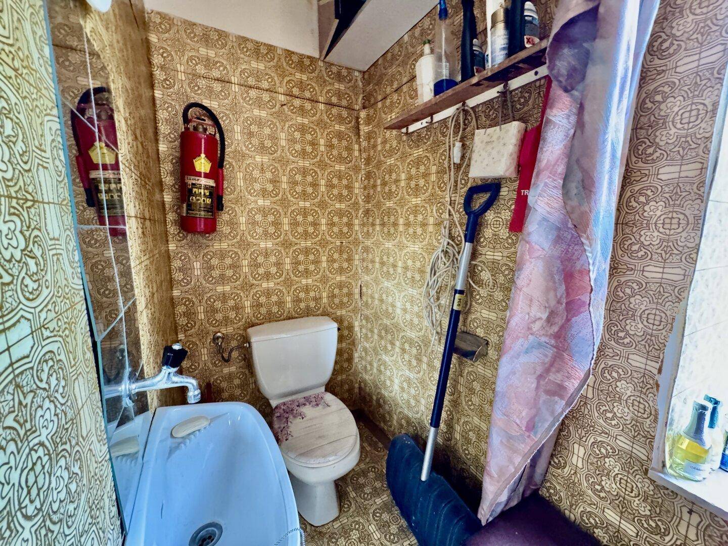Kleine Toilette mit gemusterter Tapete, WC, Waschbecken und einem Feuerlöscher an der Wand.