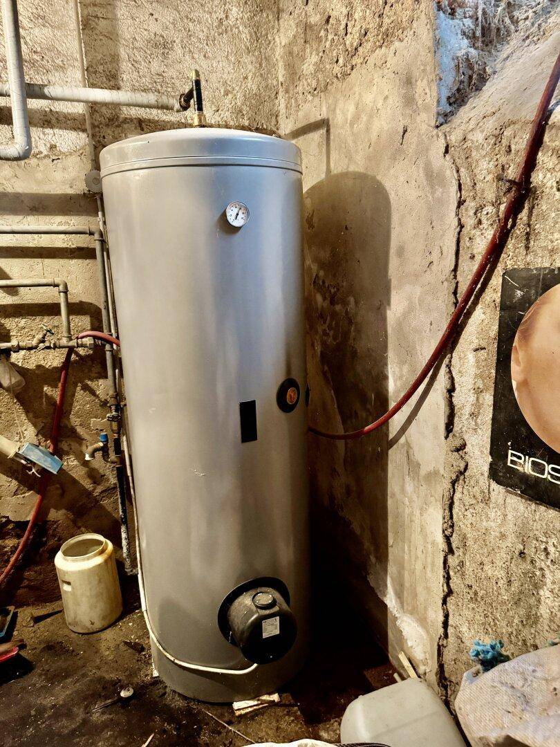 Warmwasserspeicher in einem Kellerraum mit sichtbaren Rohren und rauen Betonwänden.