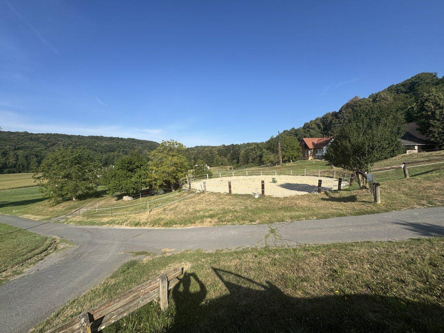 Außenreitplatz umgeben von grüner Landschaft und bewaldeten Hügeln unter blauem Himmel.