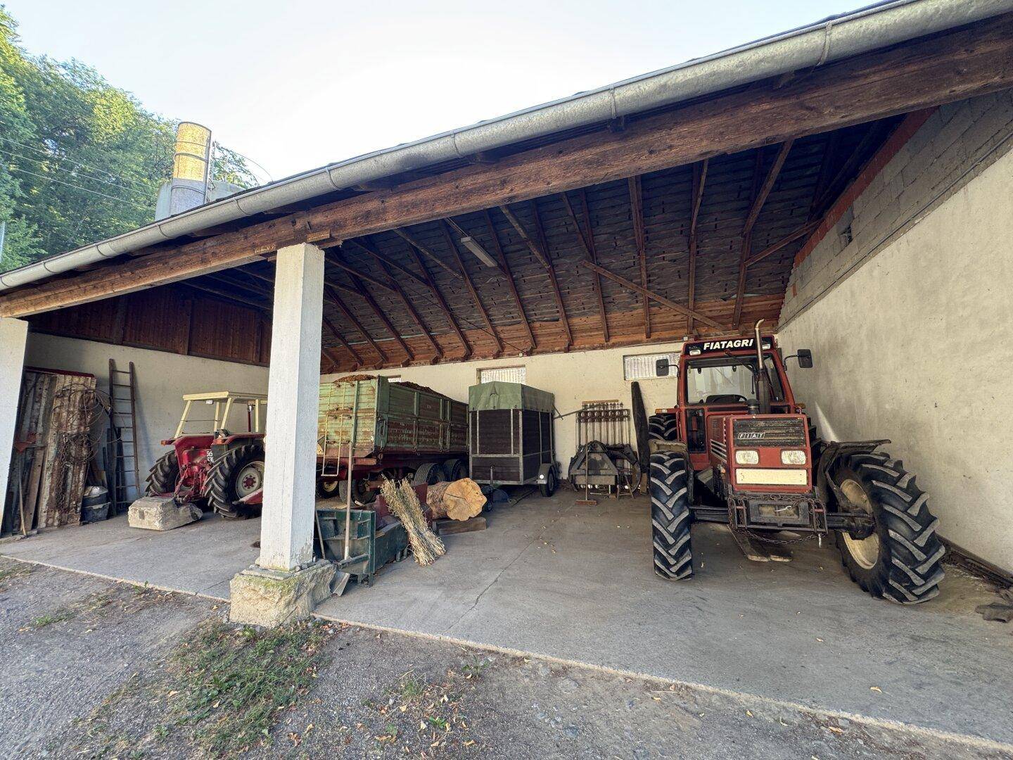 Überdachter Bereich mit landwirtschaftlichen Fahrzeugen, Holzbalken und Betonboden.