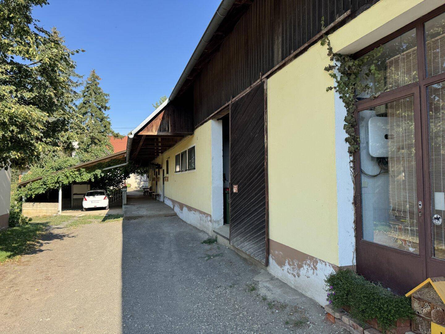 Gebäudeseite mit überdachtem Durchgang, begrüntem Eingangsbereich und Carport.