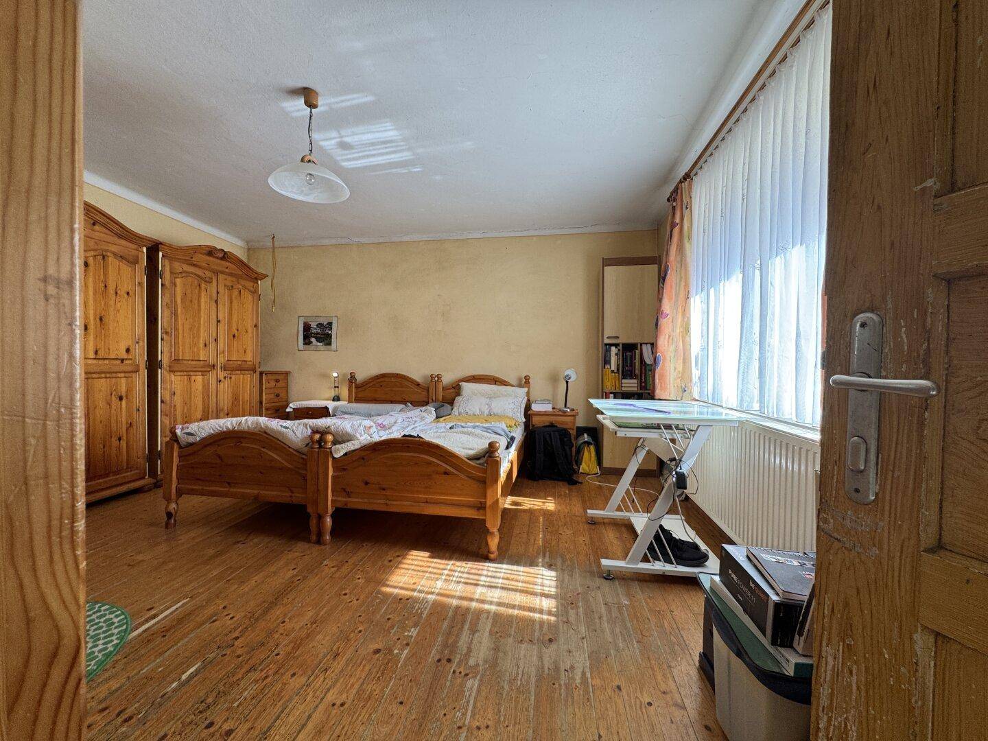 Schlafzimmer mit Holzdielenboden, Holzmöbeln und viel Tageslicht durch große Fenster.