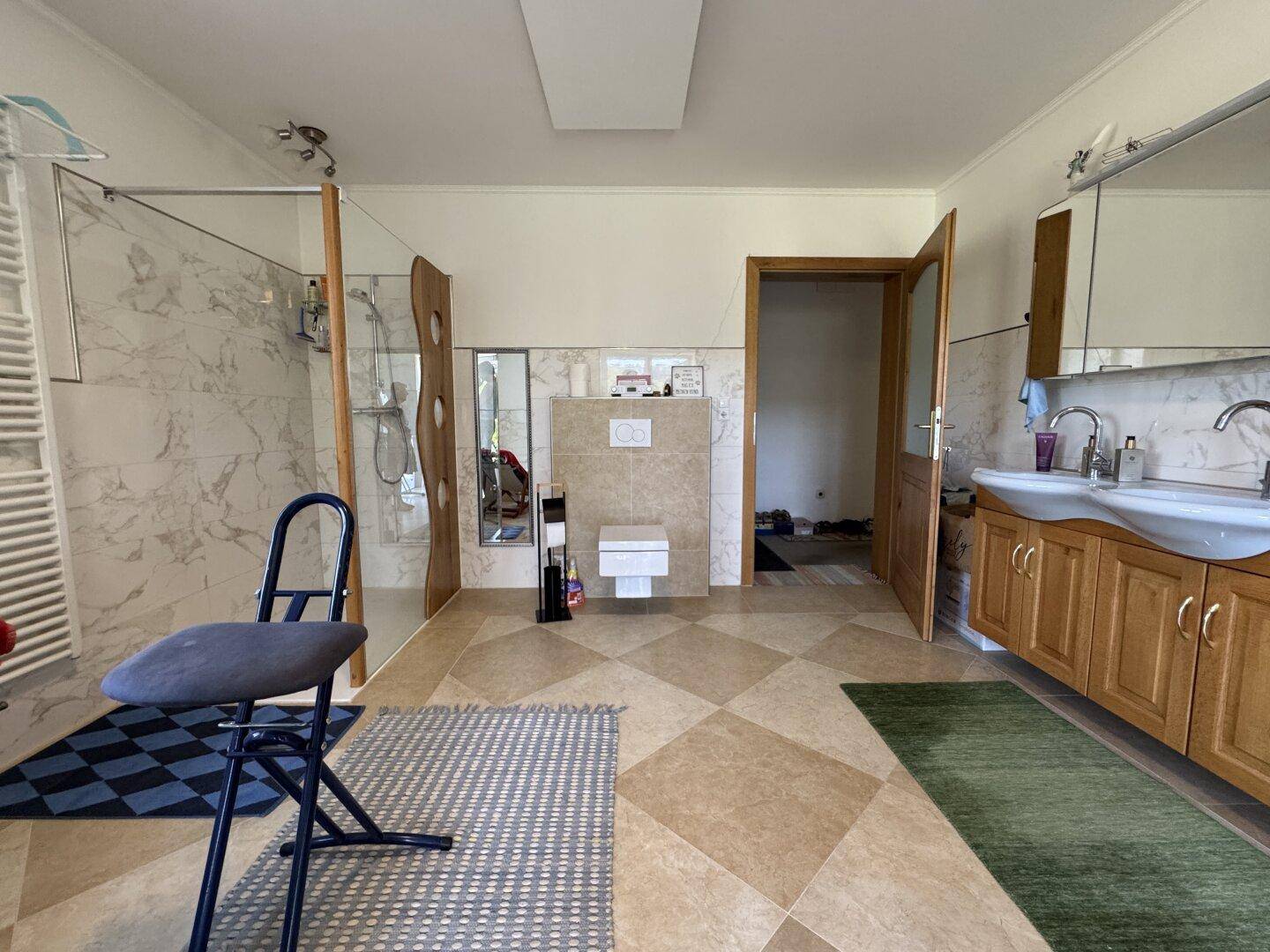 Geräumiges Badezimmer mit Doppelwaschbecken, Dusche und Fliesenboden in hellen Farbtönen.