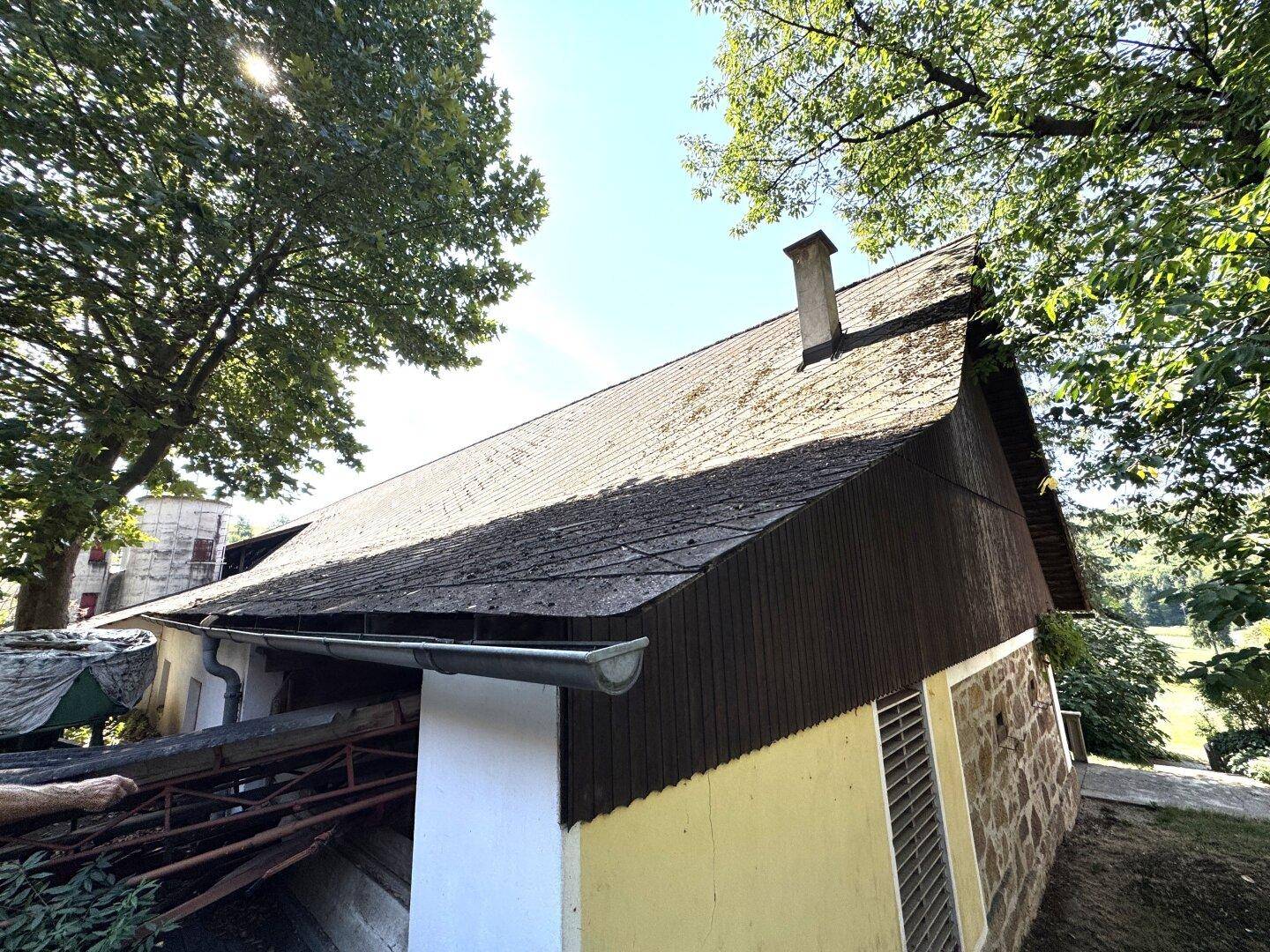 Ländliches Gebäude mit dunkler Holzfassade und Schrägdach, umgeben von hohen Bäumen an einem sonnigen Tag.