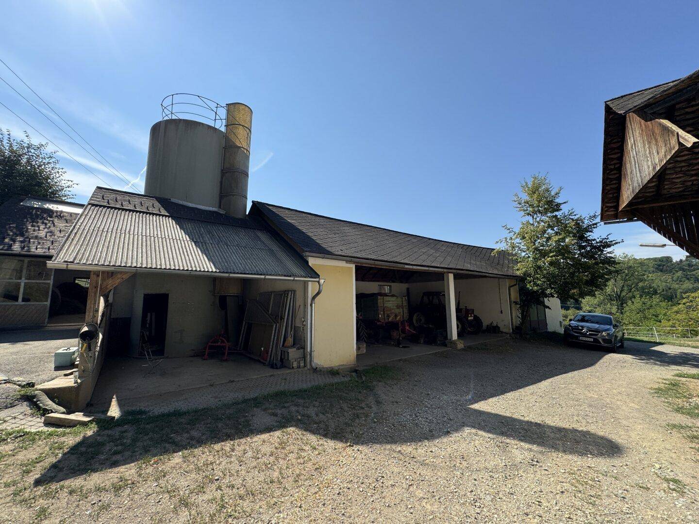 Außenansicht eines landwirtschaftlichen Gebäudes mit Silos, überdachtem Bereich und Schotterzufahrt.