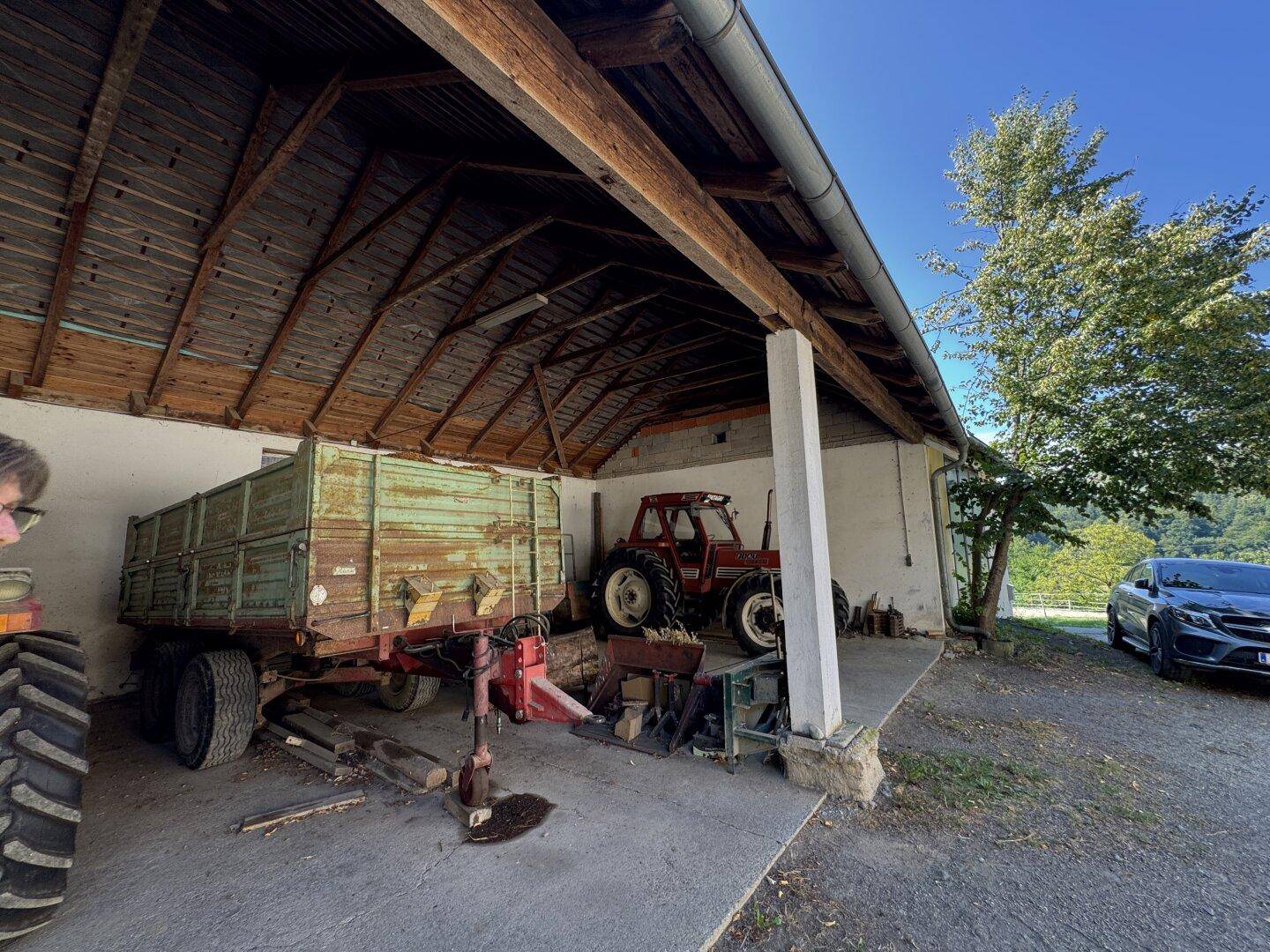 Überdachter Außenbereich mit Betonboden, landwirtschaftlichen Maschinen und einem Auto im Hintergrund.