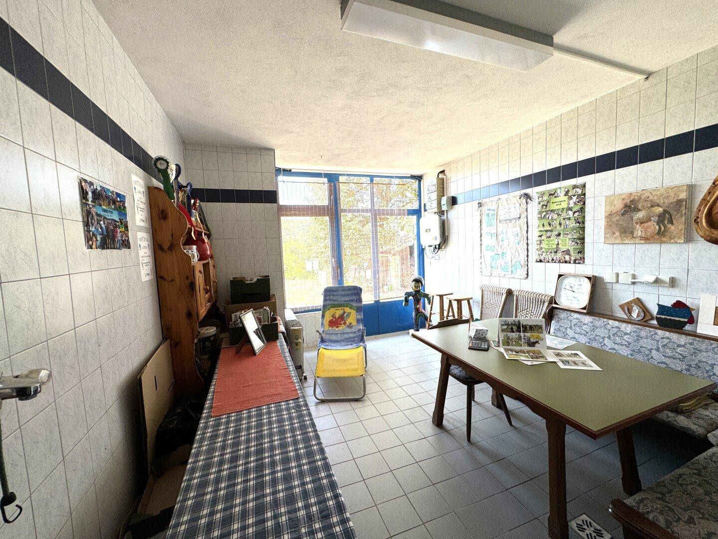 Einfacher Raum mit gefliesten Wänden und Boden, einem Tisch und Bänken, sowie einem großen Fenster.