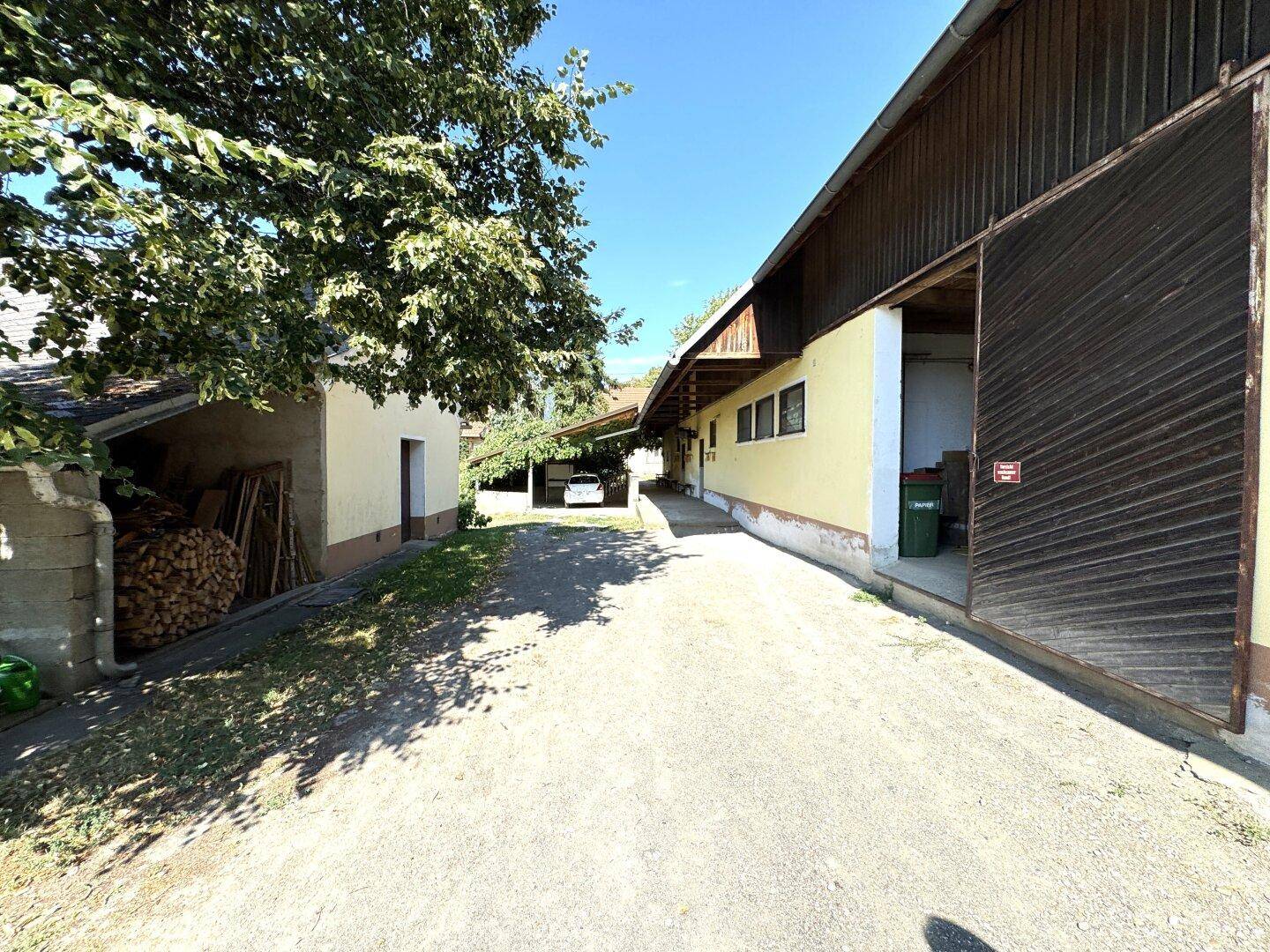 Außenansicht eines Bauernhofkomplexes mit Zufahrt, verschiedenen Gebäuden und gestapeltem Brennholz.