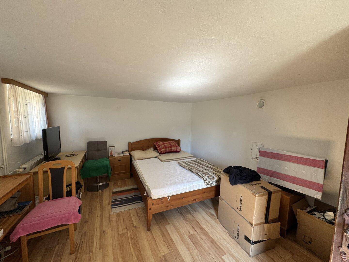 Einfaches Schlafzimmer mit Holzbett, Laminatboden und einem kleinen Fenster, funktional eingerichtet.