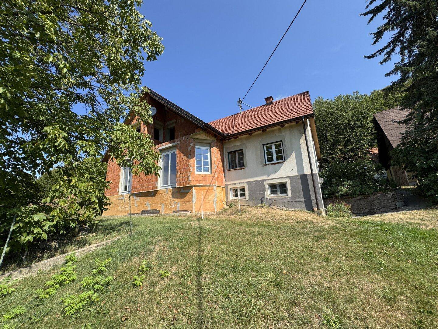Wohnhaus mit teilweise unverputzter Fassade, roten Dachziegeln und Hanglage im Grünen.