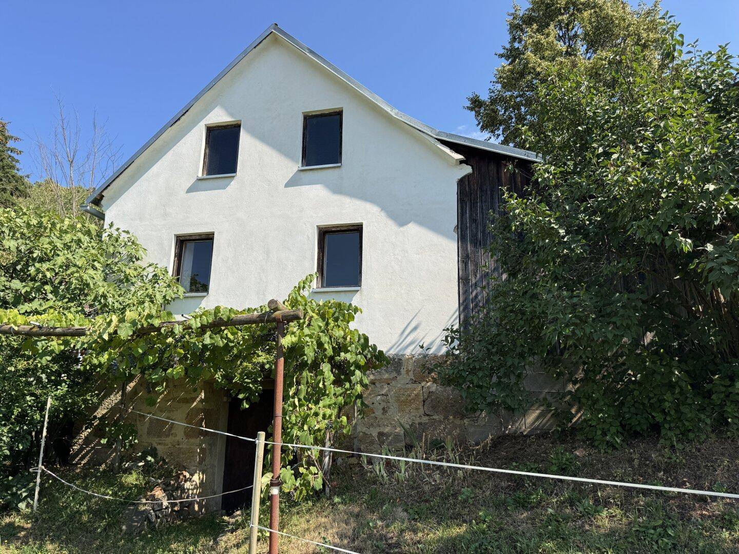 Weiß verputztes Landhaus mit Satteldach und bewachsenem Sockel, umgeben von Bäumen.