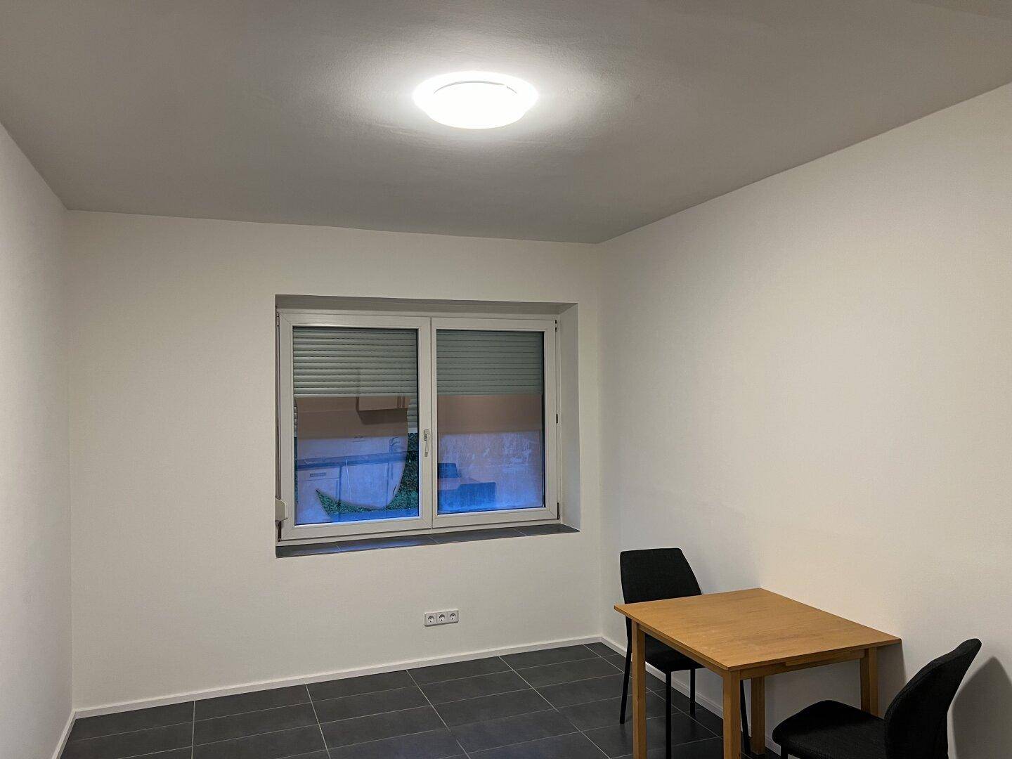 Heller Wohnbereich mit Fenster, dunklem Fliesenboden und einfacher Möblierung, ideal für ein kleines Apartment.