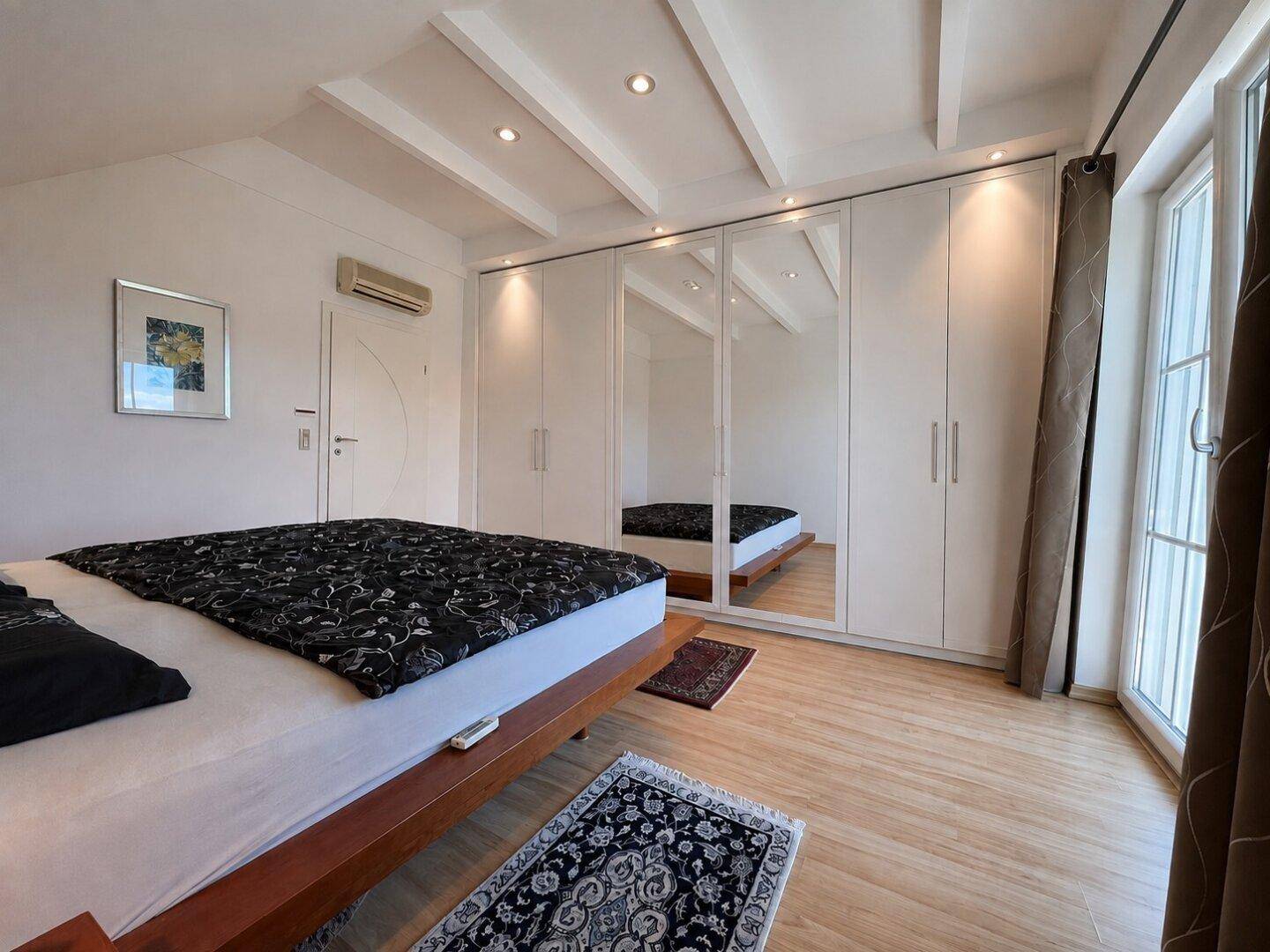 Helles Schlafzimmer mit Holzbalkendecke, Einbauschränken und Zugang zu einem französischen Balkon.