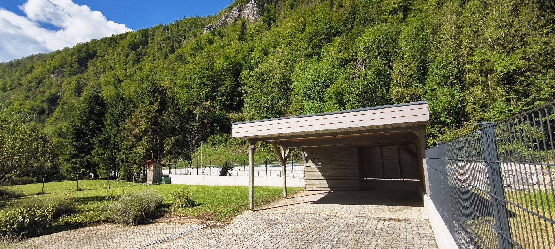 Geräumiger Carport mit gepflasterter Zufahrt, umgeben von grüner Natur und Bergen.
