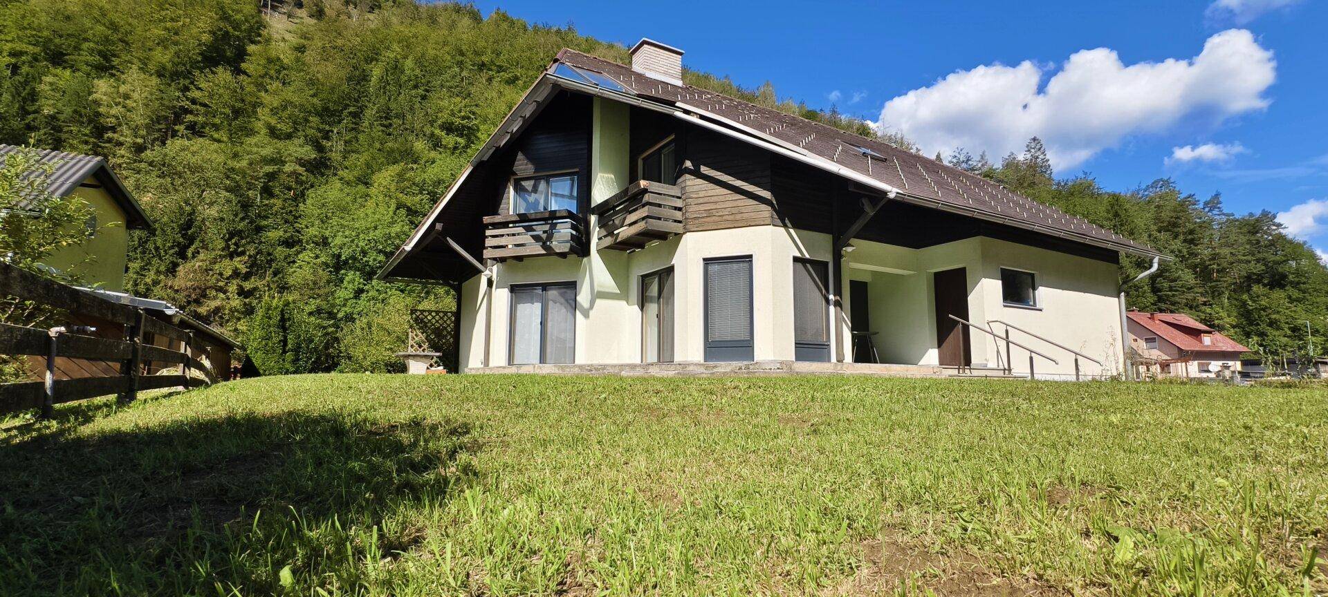 Freistehendes Haus mit Satteldach und Balkon, eingebettet in eine grüne Hügellandschaft.