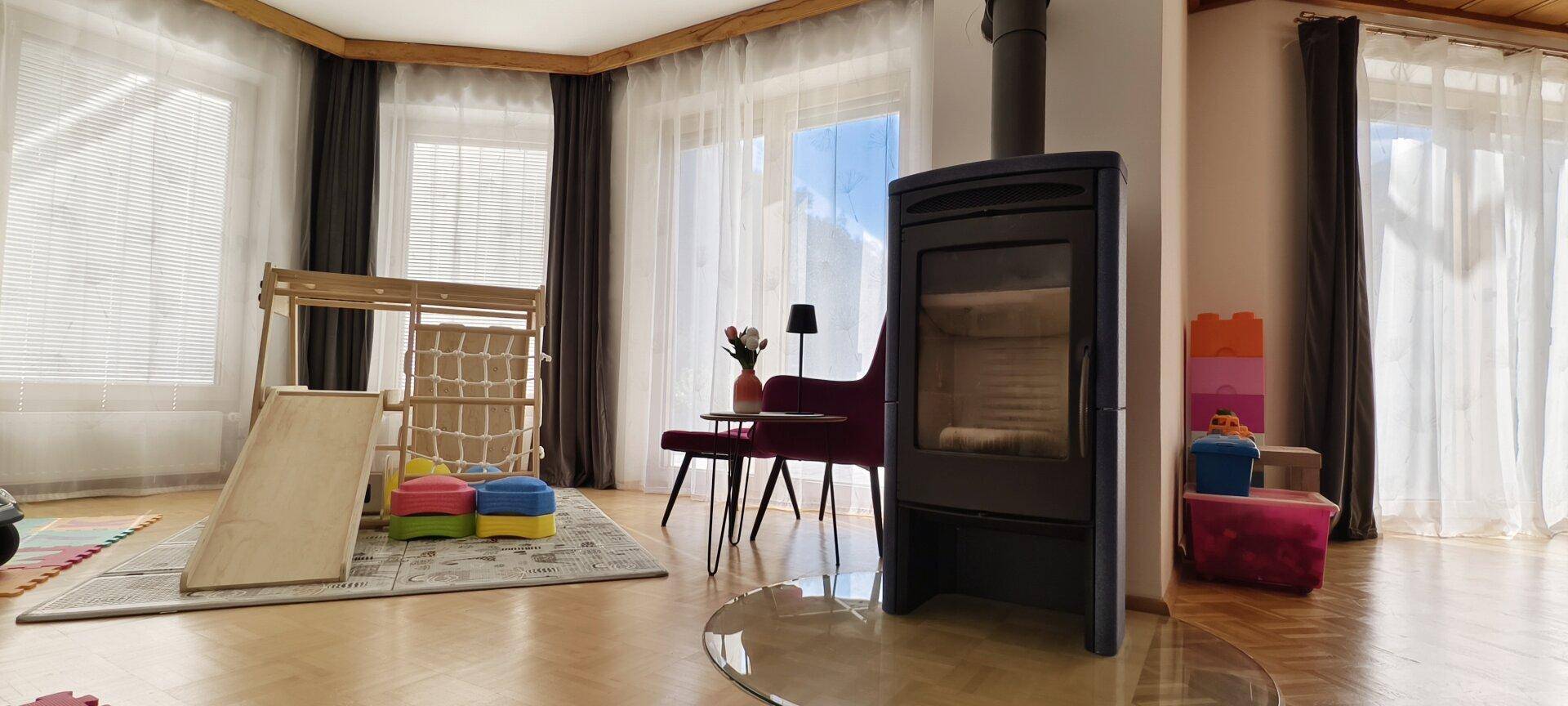 Heller Wohnbereich mit Kaminofen, großen Fenstern und Kinderspielbereich.