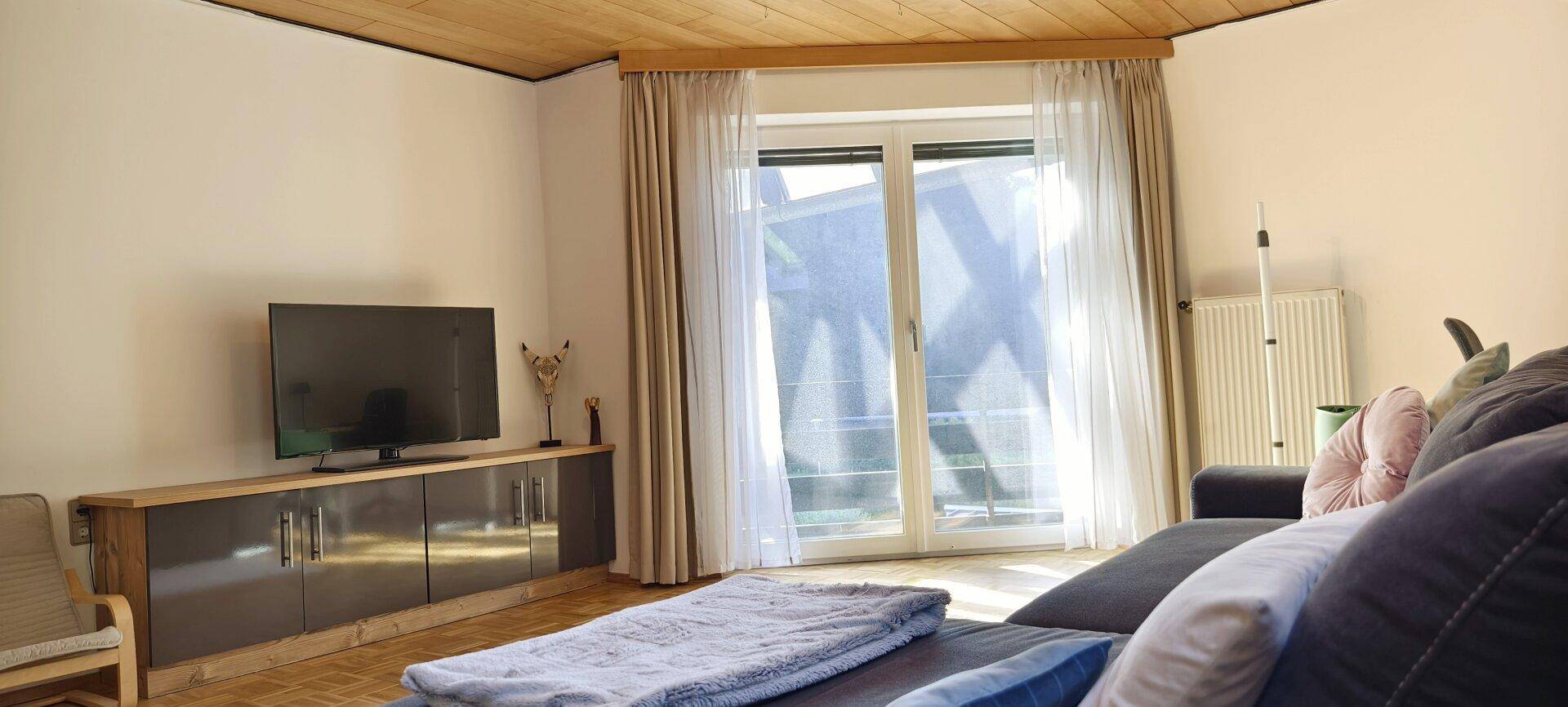 Wohnzimmer mit bequemer Couch, Fernseher und großem Fenster, das viel Tageslicht hereinlässt.