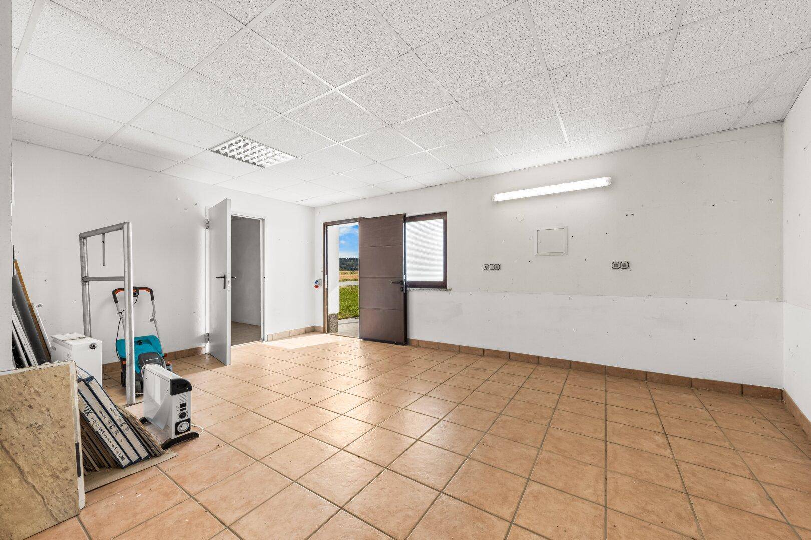 Großer, leerer Raum mit Fliesenboden, abgehängter Decke und Zugang nach draußen.