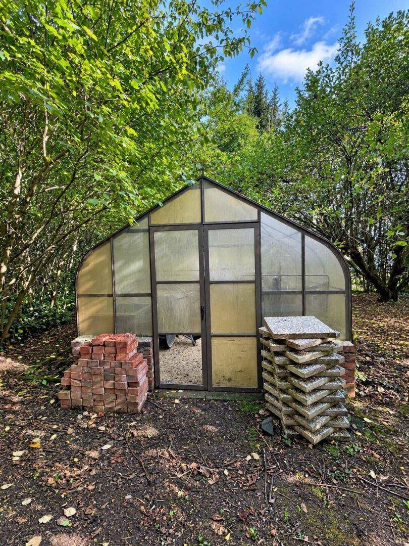 Kleines Gewächshaus im Garten, umgeben von Bäumen und Ziegelsteinen.