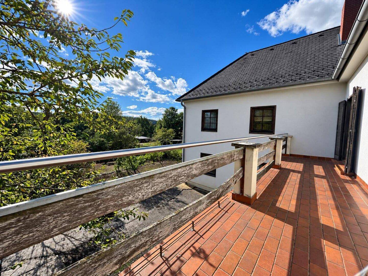 Sonnige Terrasse mit Terrakottafliesen und Holzgeländer, umgeben von Natur.