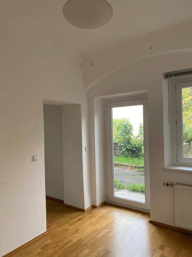 Heller Wohnraum mit Holzparkettboden und einer Tür, die ins Freie mit Grünblick führt.