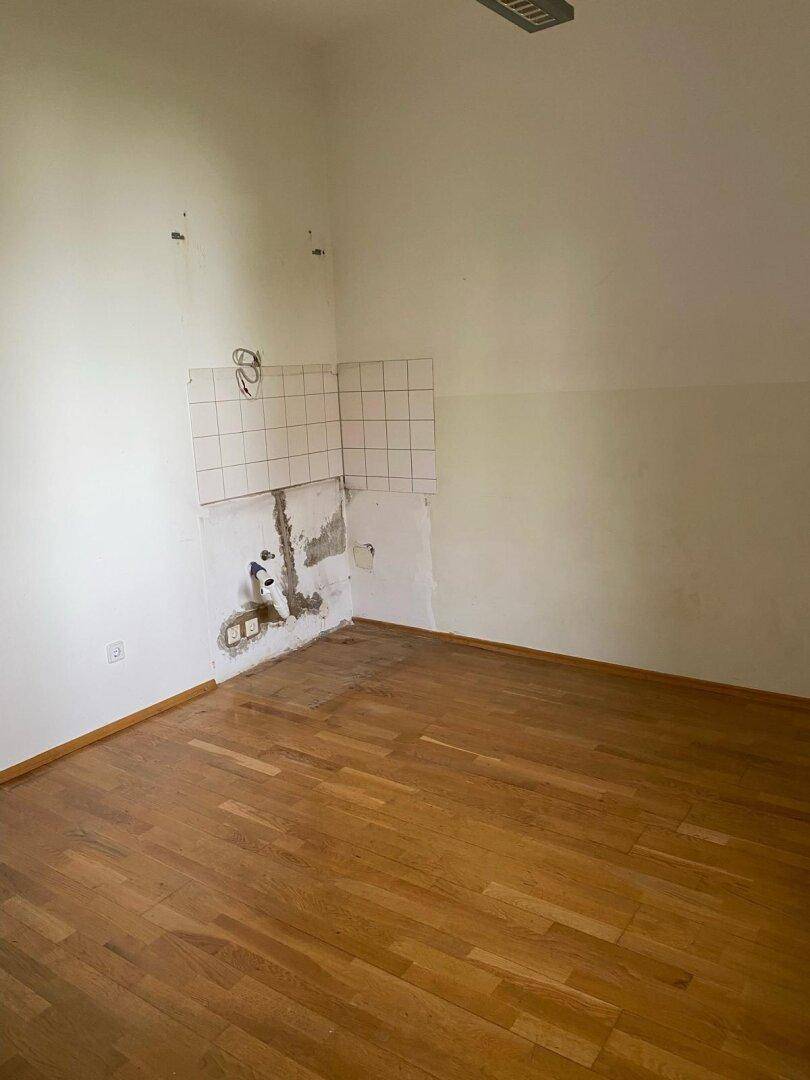 Leerer Raum mit Holzparkettboden und vorbereiteten Anschlüssen für eine zukünftige Kücheninstallation.