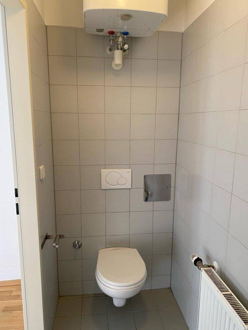 Separate Toilette mit weißen Fliesen, einem Wand-WC und einem Warmwasserboiler an der Decke.