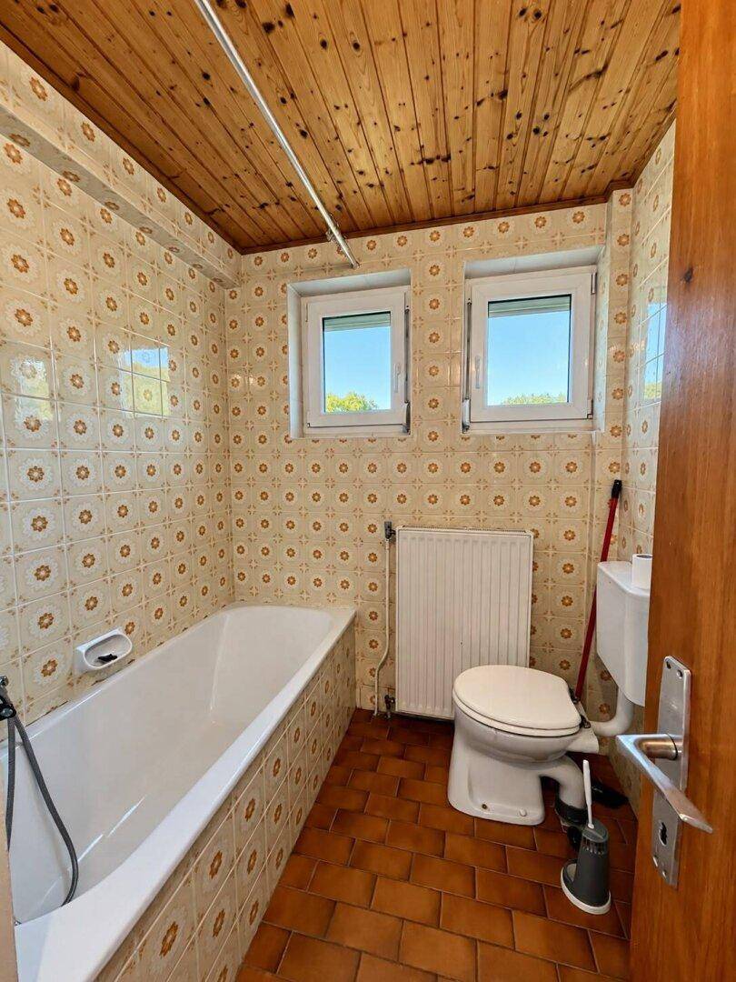 Badezimmer mit Badewanne, WC und gemusterten Fliesen, sowie Holzdecke.