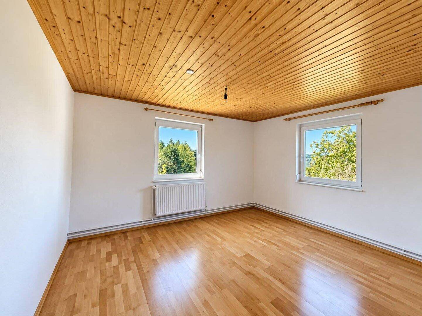Helles Zimmer mit Holzdecke und Parkettboden, zwei Fenstern und Blick ins Grüne.