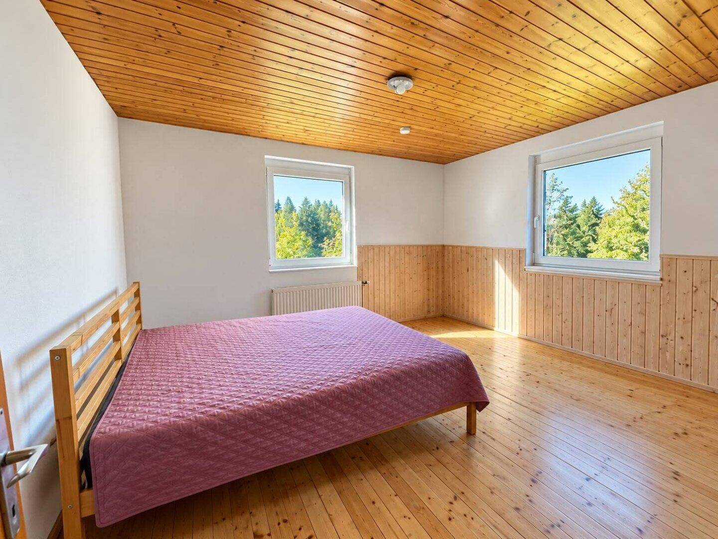 Gemütliches Schlafzimmer mit Holzdecke, Holzboden und zwei Fenstern mit Waldblick.