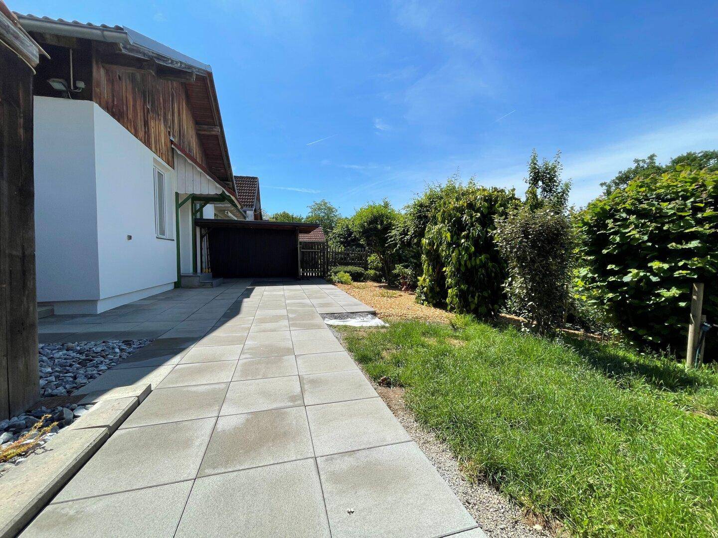 Seitenansicht des Hauses mit gepflastertem Weg, der zu einem Carport und dem grünen Garten führt.