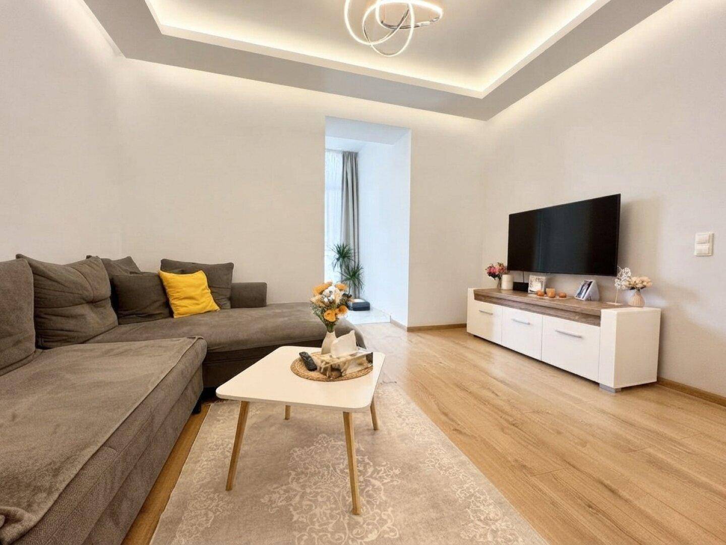 Modernes Wohnzimmer mit gemütlichem L-Sofa, Holzboden und indirekter Deckenbeleuchtung für eine angenehme Atmosphäre.