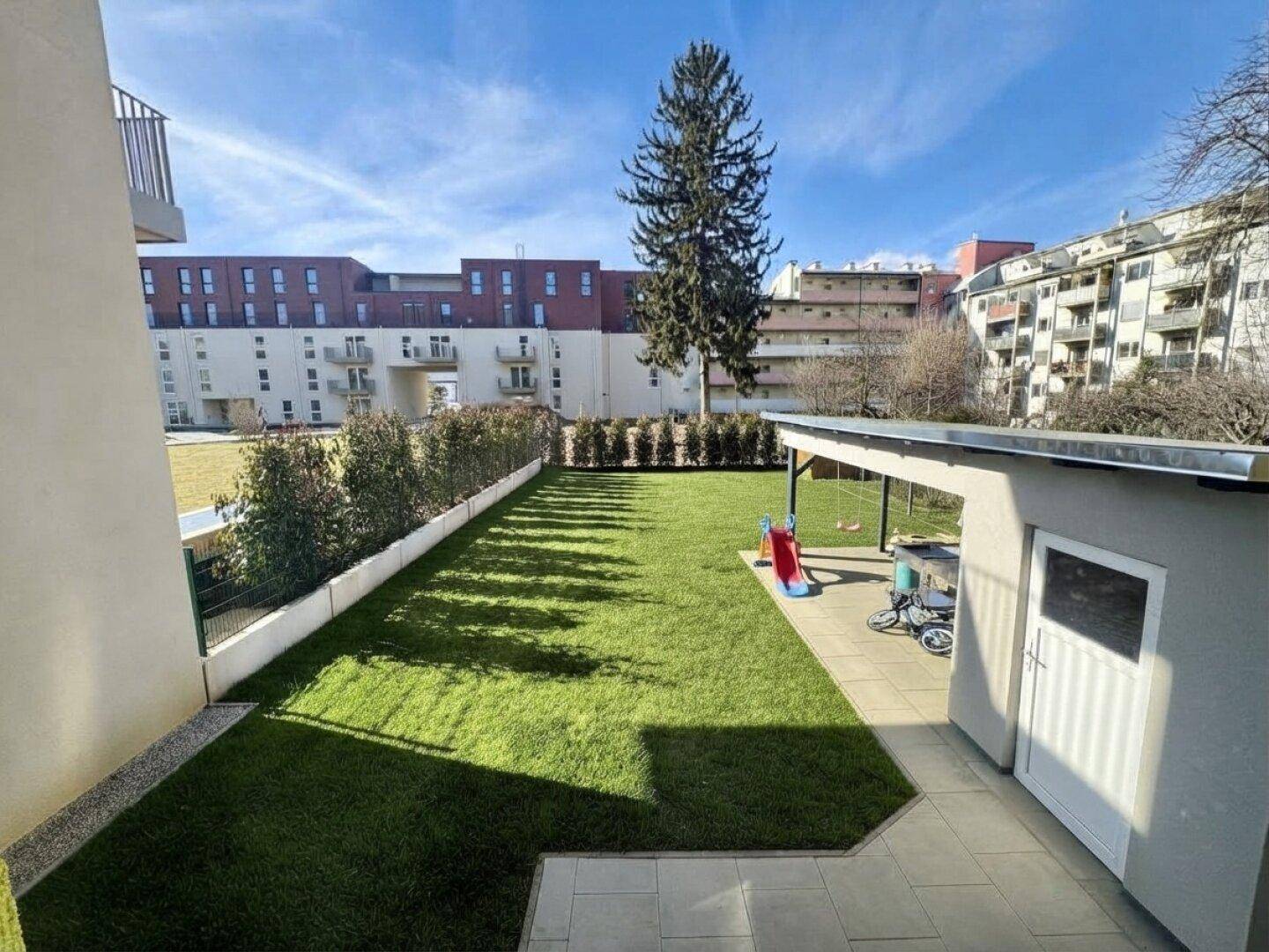 Gepflegter Gartenbereich mit Rasenfläche, überdachtem Sitzplatz und Kinderspielgeräten, umgeben von Gebäuden.
