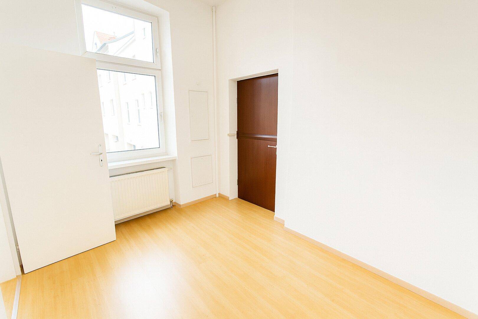 Leerer Raum mit Laminatboden, einem Fenster und einer dunklen Holztür, geeignet als kleines Büro.