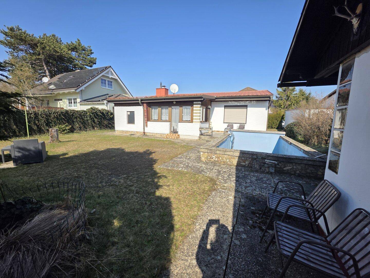 Gartenbereich mit einem Pool, einer Terrasse und Sitzgelegenheiten neben dem Haus unter strahlendem Sonnenschein.