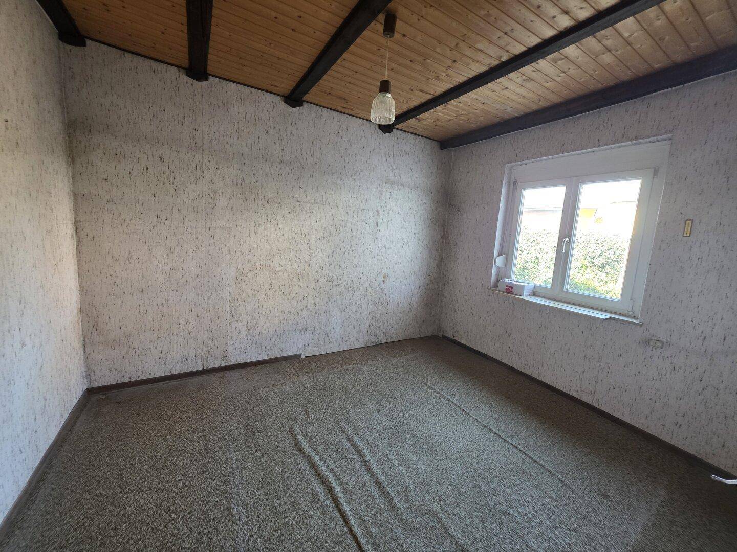 Leerer Raum mit gemusterter Tapete, Holzbalkendecke und einem Fenster mit Blick ins Grüne.