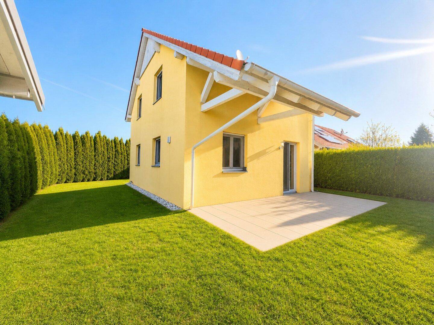 Gelbes Einfamilienhaus mit rotem Dach und einer neu angelegten Terrasse im grünen Garten.
