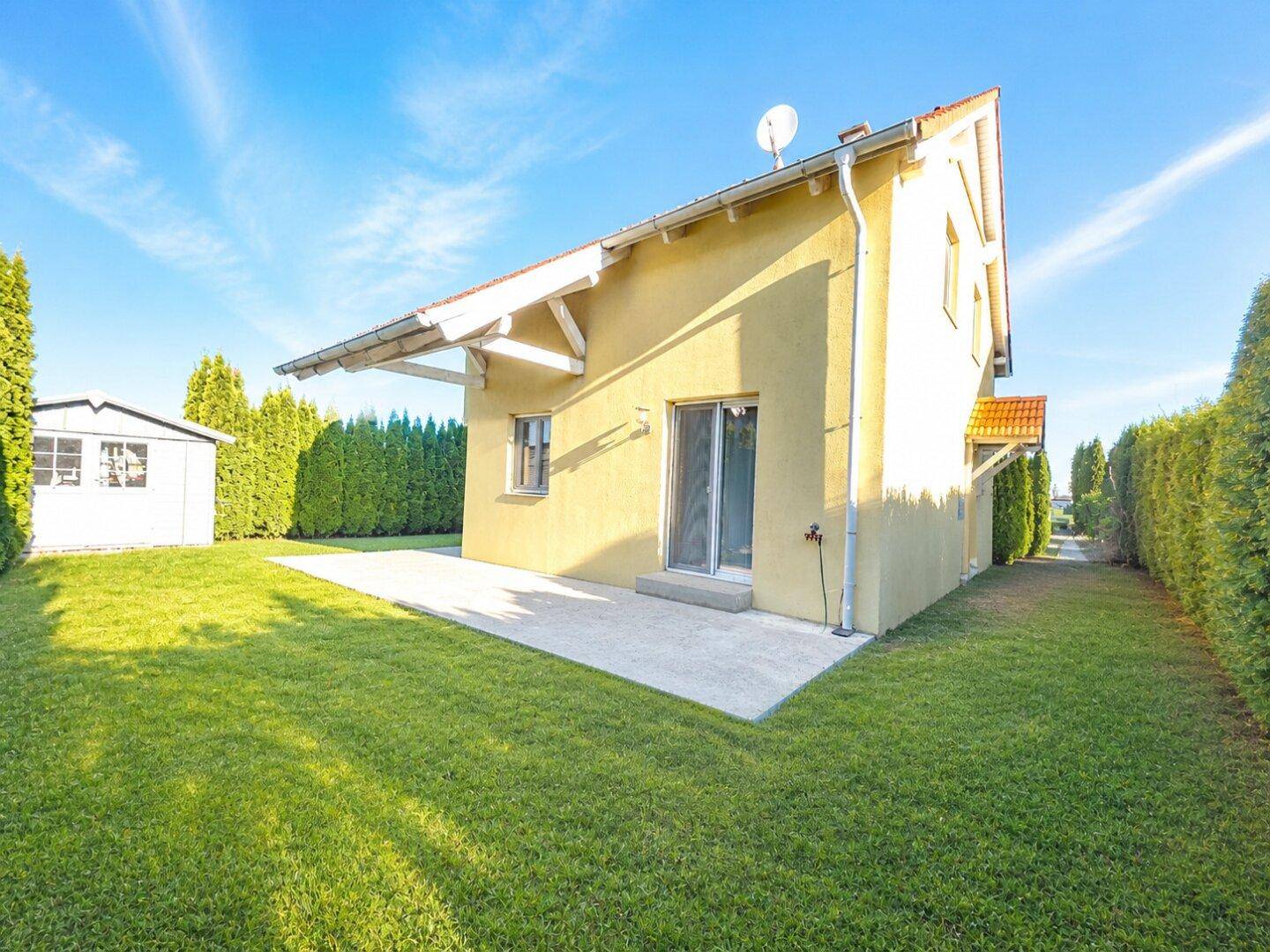 Rückseite des gelben Einfamilienhauses mit Terrasse, Garten und einem kleinen Gartenhaus.