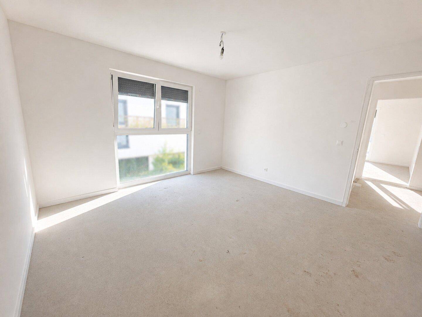 Leerer Raum im Rohbauzustand mit großem Fenster und viel Tageslicht.