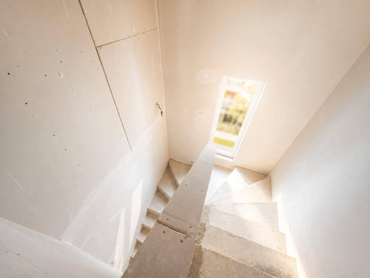 Treppenhaus im Rohbau mit Betonstufen und einem Dachfenster, das Tageslicht spendet.
