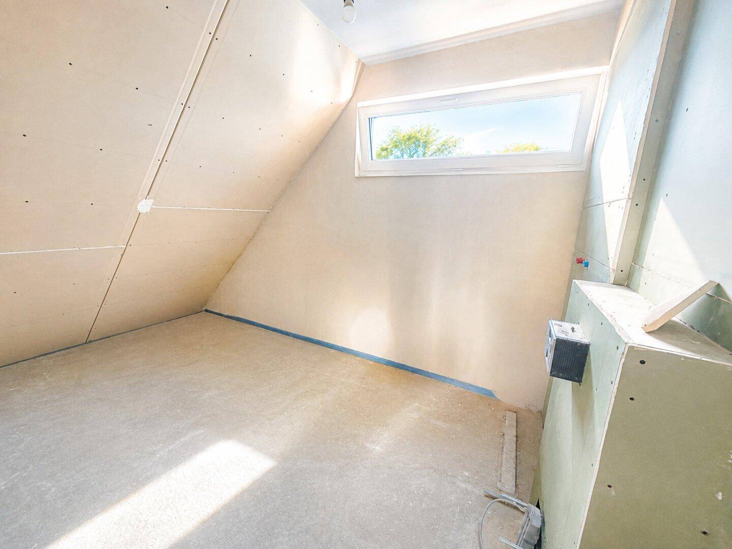 Dachgeschossraum im Rohbau mit schräger Decke und Dachfenster, das viel Licht hereinlässt.