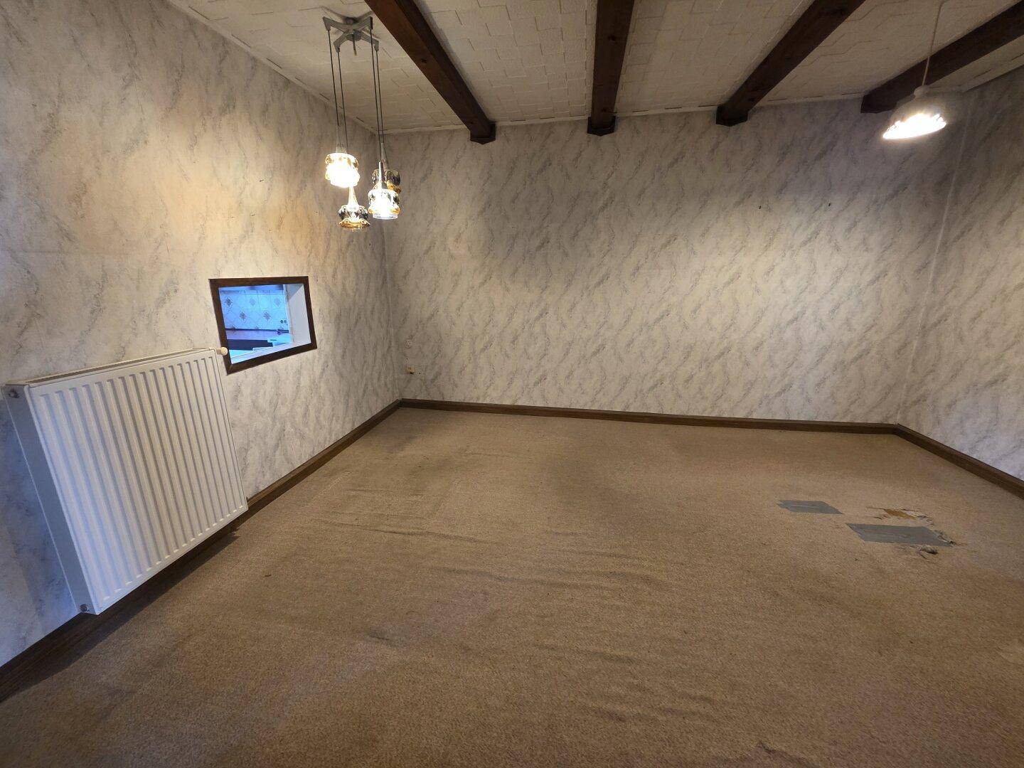 Leerer Raum mit gemusterter Tapete, Holzbalkendecke, Heizkörper und einem kleinen Fenster.