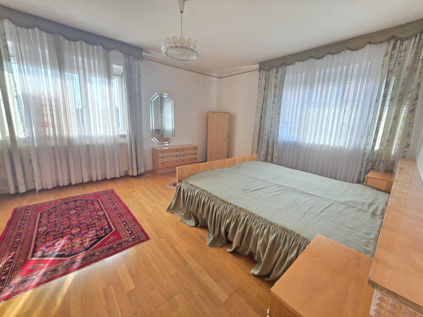 Schlafzimmer mit zwei Fenstern, Holzparkettboden und einem großen Doppelbett.